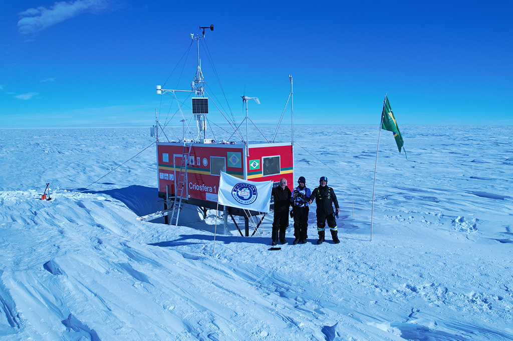 Fotografia da estação da missão criosfera ao lado de três cientistas segurando bandeira da missão.