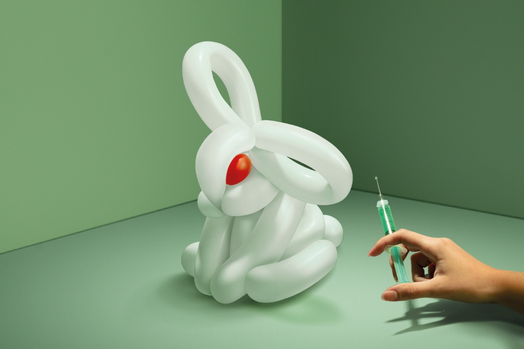 Imagem, em fundo verde, de um coelho feito com bexigas. Ao lado, uma mão feminina está segurando uma seringa com agulha e conteúdo verde.