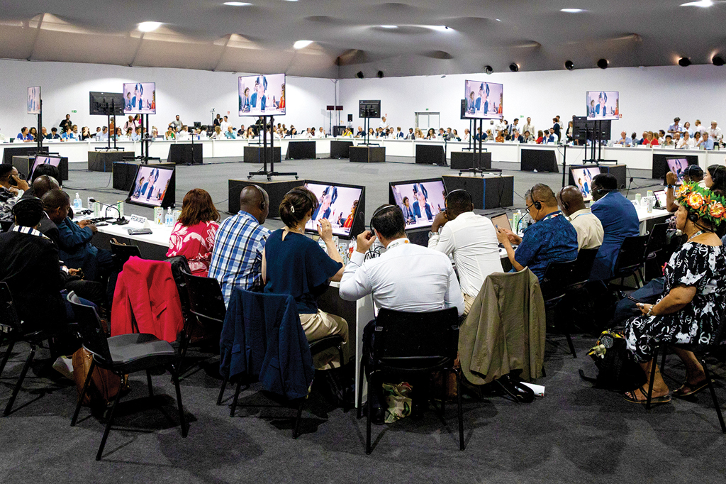 Imagem de pessoas reunidas, numa enorme sala, estão sentadas, assistindo uma reunião on-line.