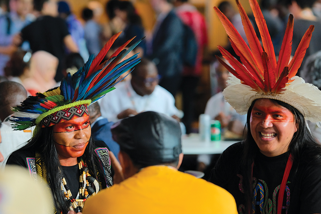 Imagem de três homens conversando. Dois deles estão usando cocar.