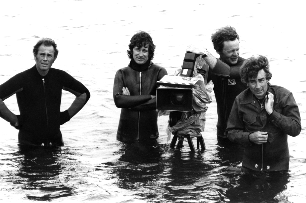 Imagem de quatro homens com água até a cintura e equipamento de filmagem.