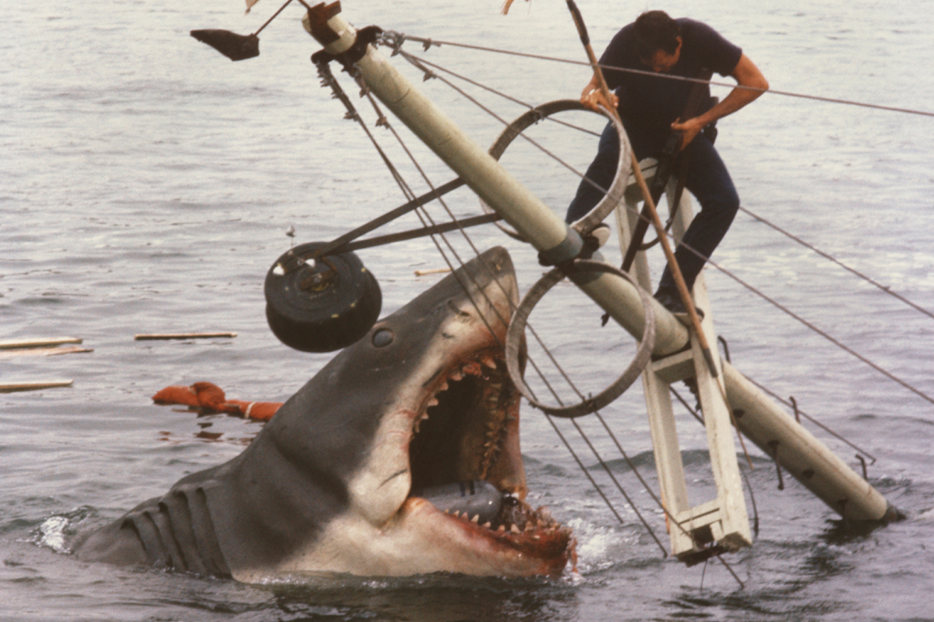 Imagem de um homem dependurado no mastro de barco afundando enfrentando um enorme tubarão. Cena do filme