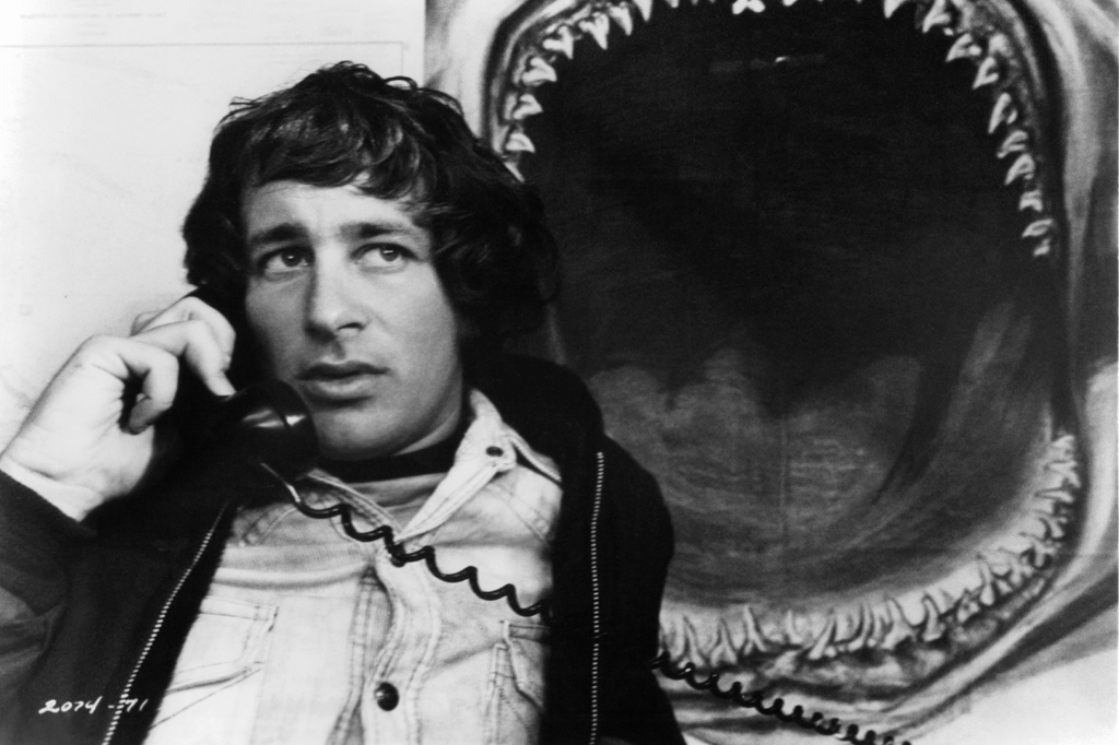 Imagem de um jovem falando ao telefone. Ao fundo, vê-se um cartaz de um tubarnão com a boca aberta.