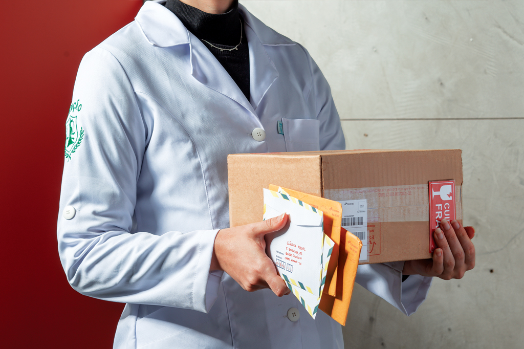 Imagem de uma mulher, vestindo jaleco branco, carregando uma caixa de papelão e alguns envelopes.