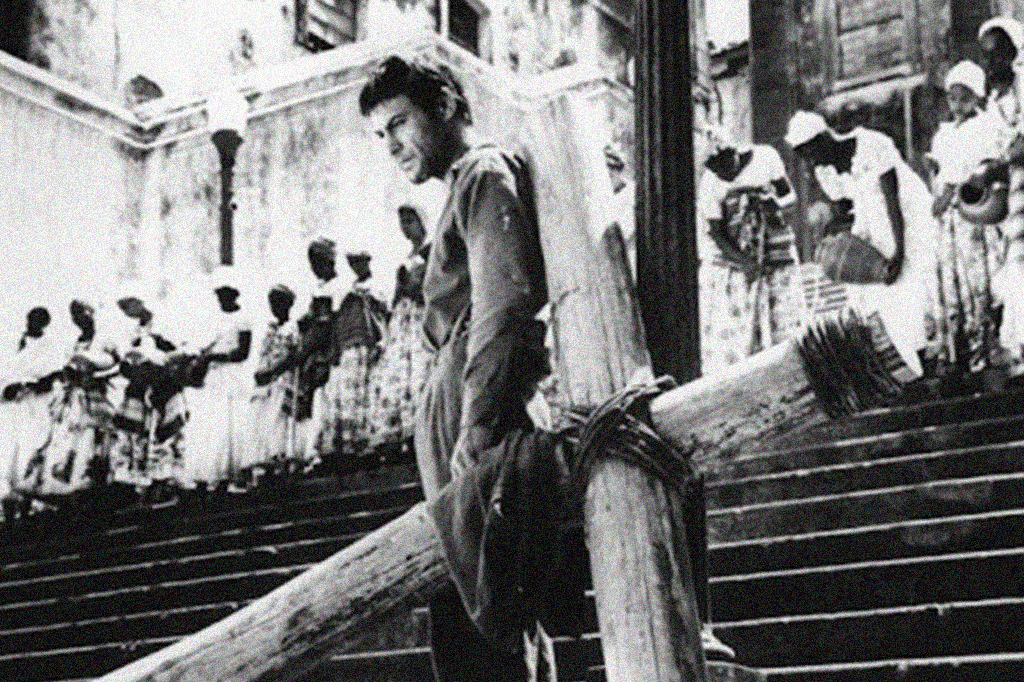 Zé Burro ao lado de uma cruz de madeira e de frente para uma escadaria