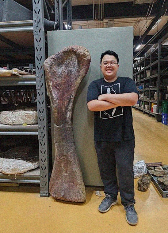 Homem asiático sorridente, de óculos e camiseta preta, braços cruzados, posa ao lado de um enorme osso fóssil marrom, em um laboratório com prateleiras cheias de amostras