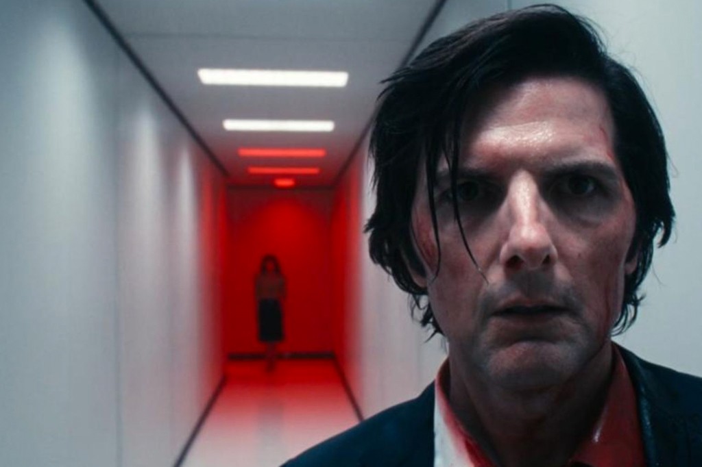 Homem de cabelo escuro e rosto sujo de sangue, com expressão de choque, olhando para frente. Ao fundo, um corredor iluminado em vermelho com uma figura feminina ameaçadora distante.