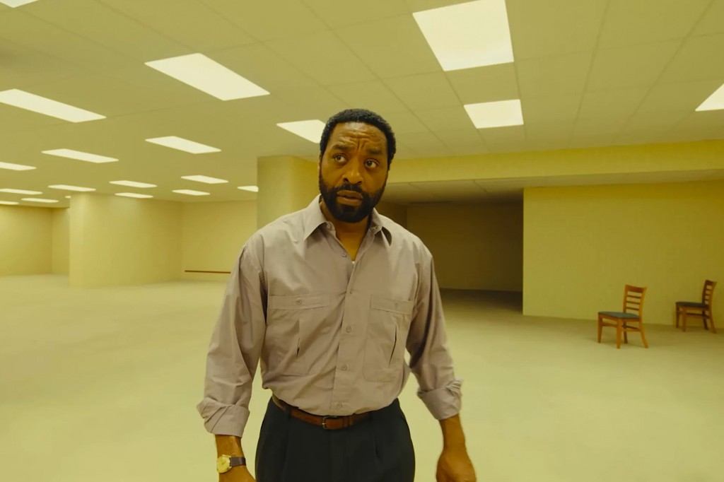 Homem negro de barba, camisa cinza e calça escura, com expressão confusa, em um ambiente vazio de paredes e chão amarelos, com luzes retangulares no teto e duas cadeiras de madeira ao fundo.