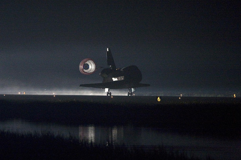 Ônibus espacial escuro pousando em pista iluminada à noite, com paraquedas de freio aberto e reflexo na água à frente