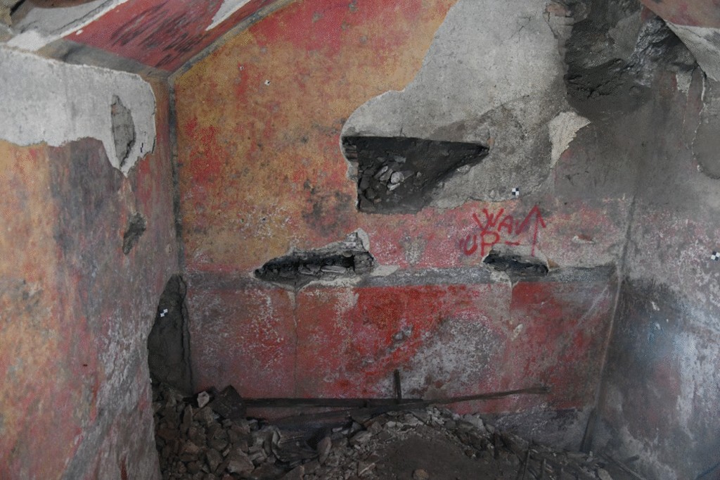 Interior de uma estrutura antiga com paredes vermelhas e amarelas descascadas, revelando concreto cinza e buracos. No chão, há entulho de pedras e escombros. A parede central exibe a pichação WAK UP em vermelho