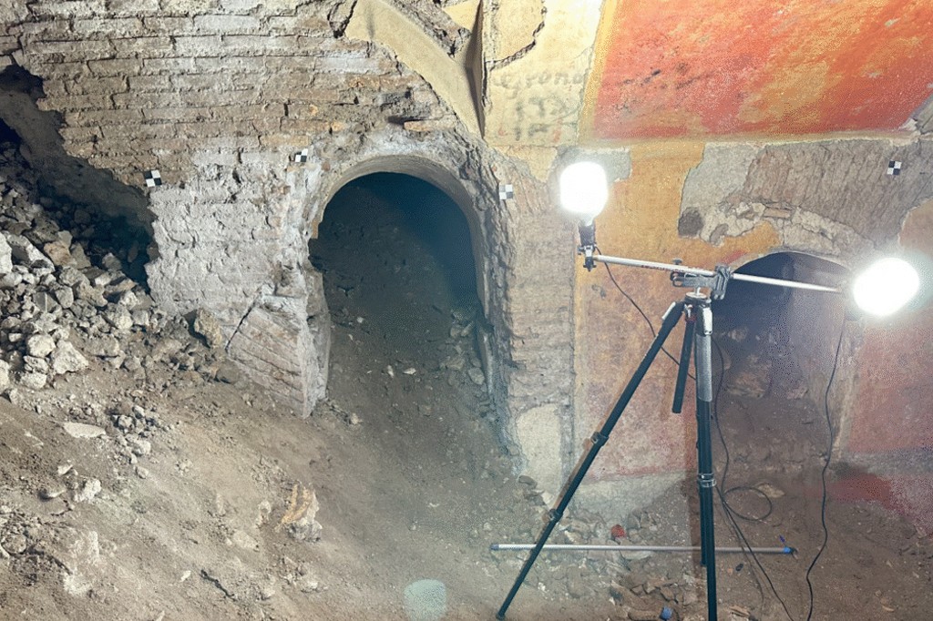 Interior de uma escavação arqueológica com duas aberturas em arco na parede de tijolos e terra, iluminadas por um tripé com duas luzes fortes. O chão é de terra e pedras soltas, e a parede superior tem vestígios de pintura avermelhada e amarelada
