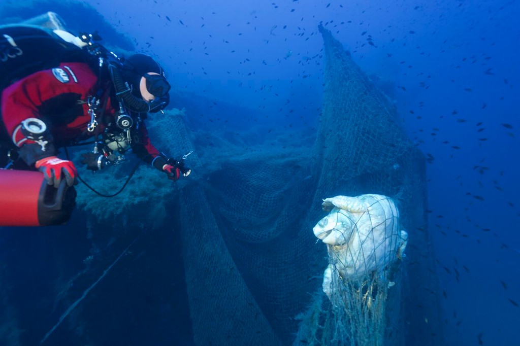 Mergulhador em roupa vermelha e preta observa uma tartaruga marinha presa em uma rede de pesca fantasma no fundo do oceano azul escuro, com pequenos peixes ao redor