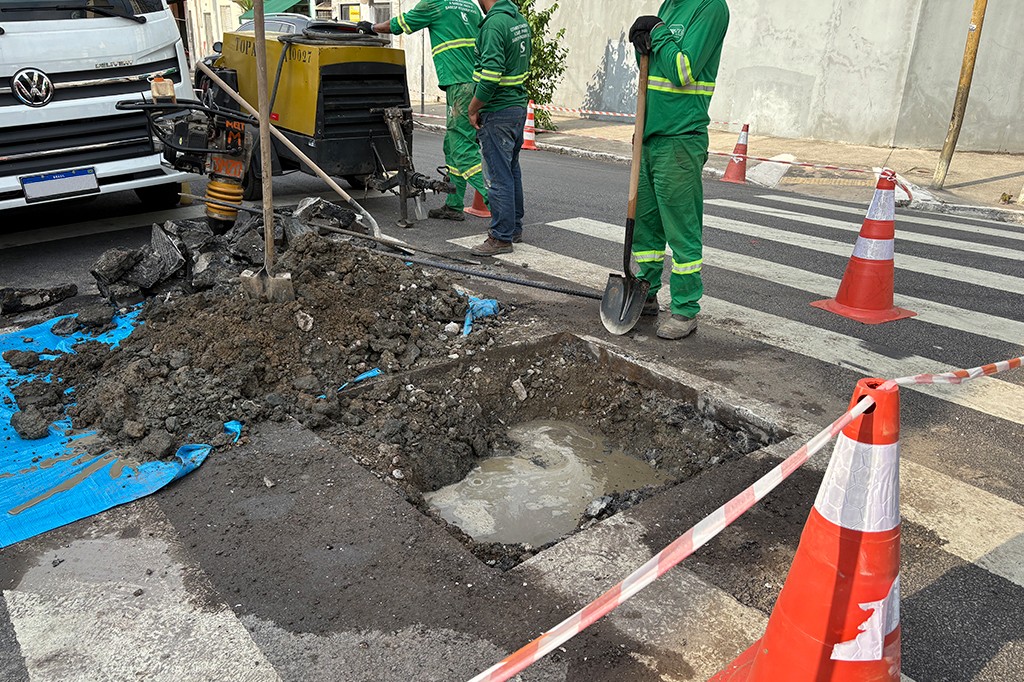 Três trabalhadores de uniforme verde operam equipamentos e uma pá em uma obra de rua, com um buraco cheio de água e terra, cones de sinalização e uma faixa de pedestres ao fundo