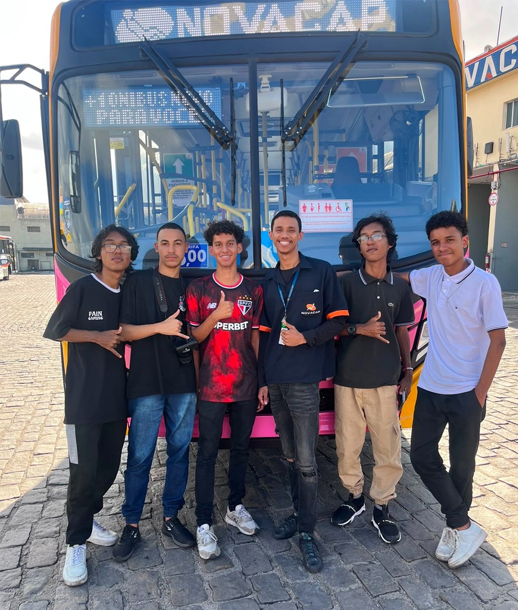 Seis jovens sorridentes, com idades entre 15 e 25 anos, posam em frente a um ônibus rosa e amarelo da empresa Novacap. O ônibus exibe no letreiro Novacap e Temos um lugar para você. Os jovens vestem roupas casuais, incluindo camisetas de times de futebol e uma camisa polo. O chão é de paralelepípedos.