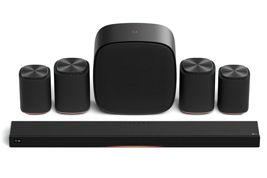 Sistema de som surround preto com soundbar retangular na frente, subwoofer central e quatro caixas de som cilíndricas ao redor, todos com detalhes em cobre na base.
