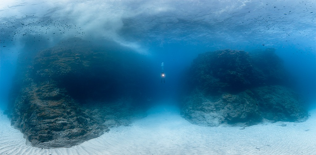 Mergulhador solitário com lanterna ilumina o fundo do mar azul profundo, entre duas grandes formações rochosas escuras e um leito de areia clara. Pequenos peixes nadam perto da superfície.