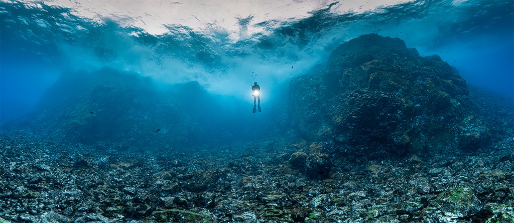 Mergulhador solitário com uma lanterna subaquática brilhante, nadando em águas azuis profundas sobre um leito marinho rochoso e irregular. A superfície da água, acima, reflete a luz do sol em ondas. À direita, uma grande formação rochosa escura se eleva do fundo.