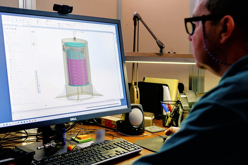 Homem de óculos e camisa azul, de costas, usando um computador Dell com um software de design 3D exibindo um cilindro transparente com partes internas azuis e roxas. A mesa tem teclado, mouse, alto-falante, telefone e porta-canetas.