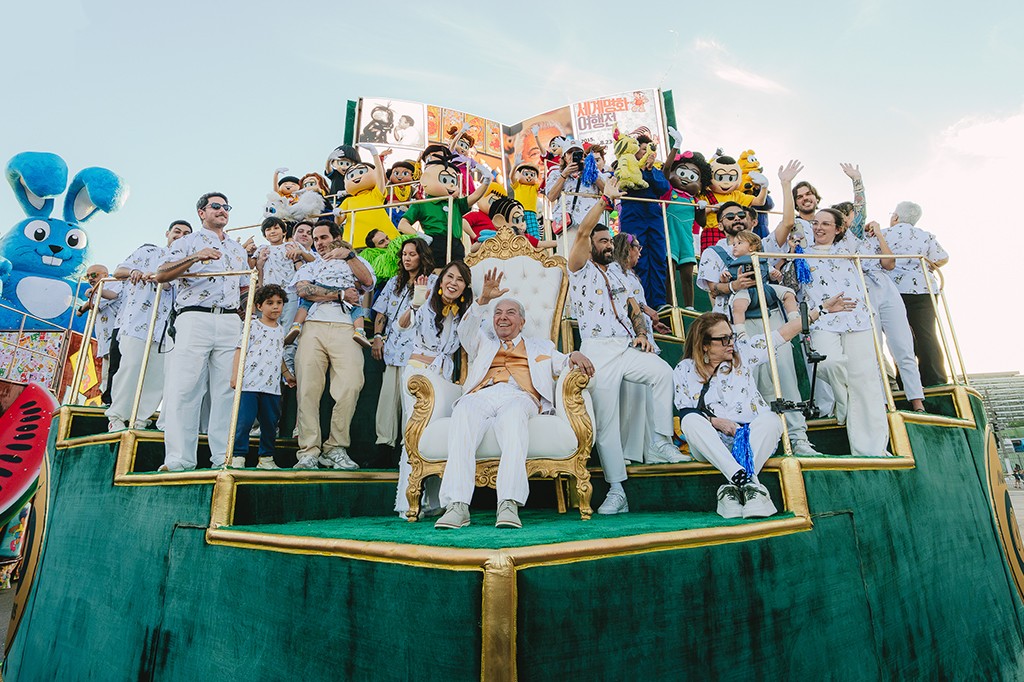 Um grupo de pessoas, incluindo crianças e adultos, em um carro alegórico verde com detalhes dourados. Um homem idoso sorridente, de terno branco e colete laranja, acena sentado em um trono branco e dourado. Outros acenam e sorriem, alguns com fantasias de personagens de desenho. Ao fundo, um livro gigante aberto com texto em coreano e um coelho azul inflável. O céu está claro