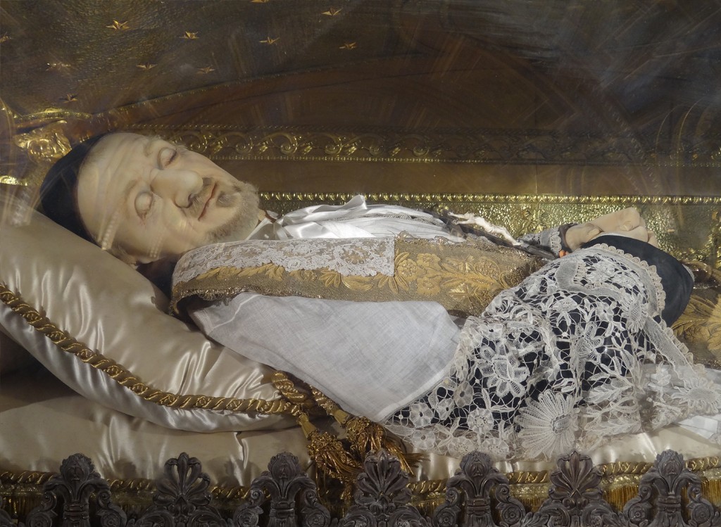 Corpo de São Vicente de Paulo, com barba grisalha e olhos fechados, deitado em um caixão dourado com detalhes ornamentados. Ele veste uma túnica branca e preta com rendas e bordados dourados, sobre um travesseiro de seda creme