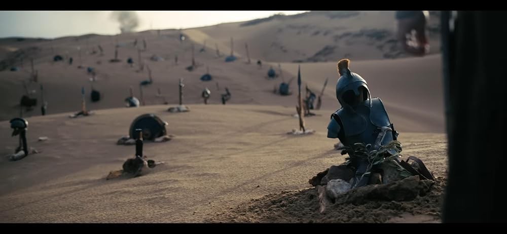 Armadura antiga de guerreiro, com capacete e crista laranja, semi-enterrada na areia de um deserto. Ao fundo, mais armaduras e armas espetadas na areia, e fumaça no horizonte.