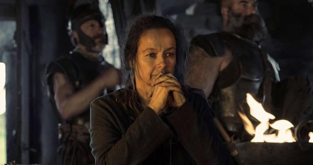 Mulher de meia-idade com cabelos escuros e expressão preocupada, mãos unidas, vestindo roupa escura. Ao fundo, dois homens barbudos em armaduras e uma fogueira acesa no canto inferior direito, em ambiente escuro
