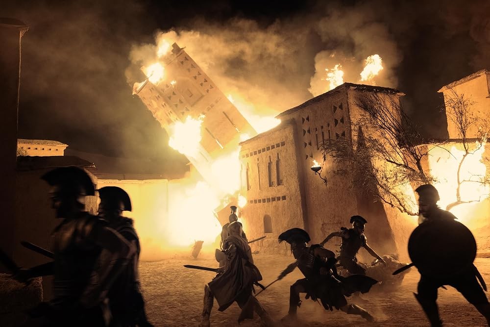 Guerreiros em silhueta correm em meio a um cenário noturno de destruição, com edifícios antigos em chamas e fumaça densa subindo ao céu