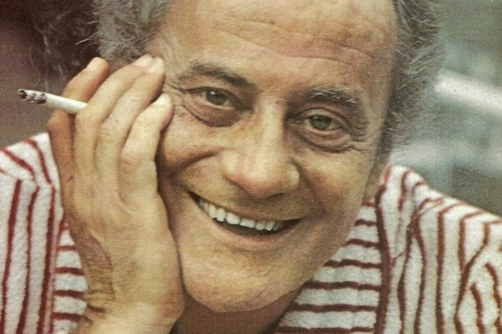 Um homem de pele clara, cabelos grisalhos e rosto enrugado, sorri amplamente, com a mão direita apoiada na bochecha e um cigarro aceso entre os dedos. Ele veste uma camisa listrada em branco e vermelho. Retrato de Paulo Autran.