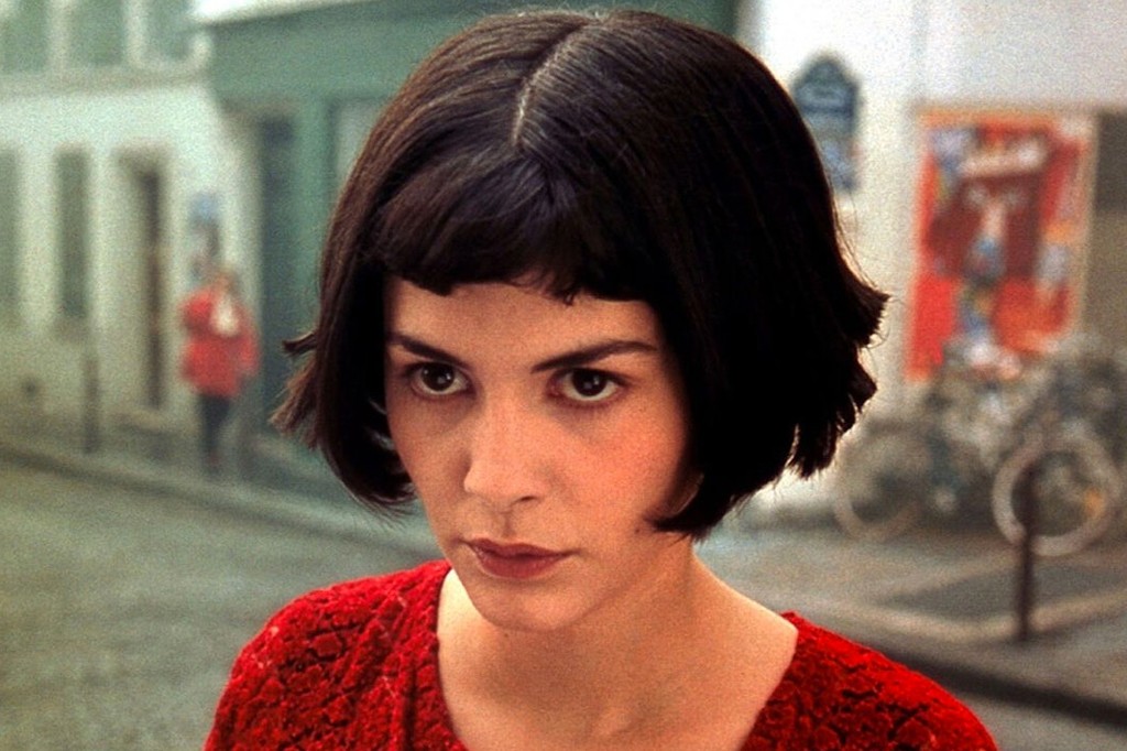 Mulher de cabelo curto e escuro, com franja reta, veste um suéter vermelho texturizado e olha diretamente para frente com expressão séria. Ao fundo, uma rua borrada com edifícios claros e uma pessoa de casaco vermelho distante.