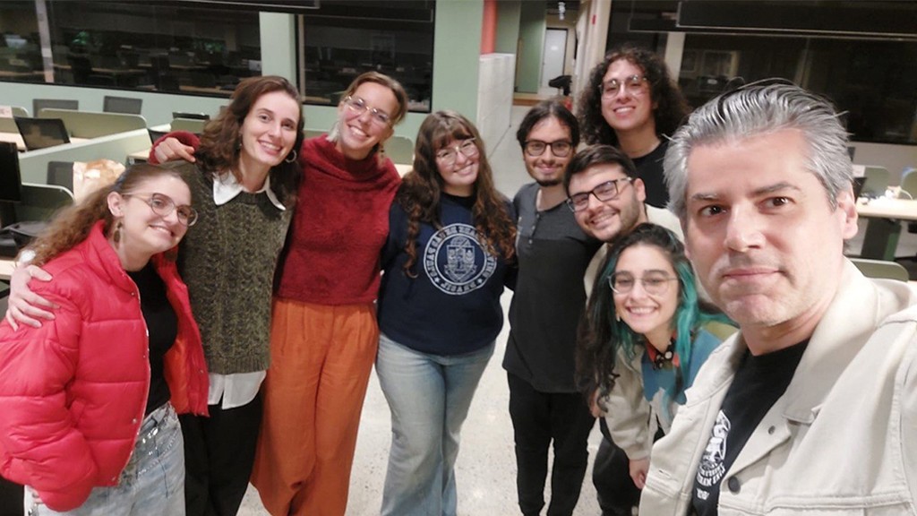 Grupo de nove pessoas sorrindo para a câmera em um ambiente interno com mesas e cadeiras ao fundo. Todos parecem felizes e descontraídos.