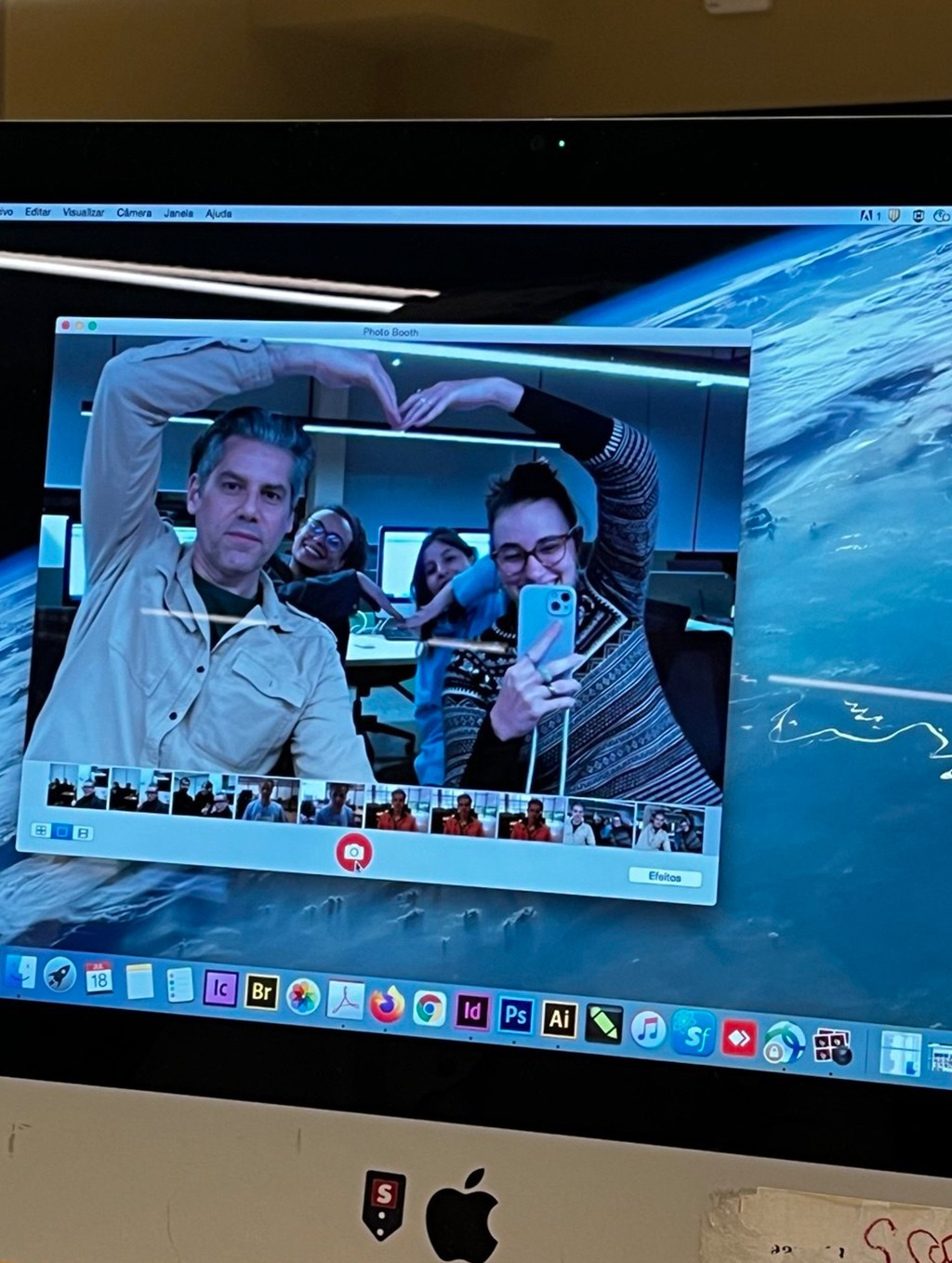Quatro pessoas sorrindo, duas à frente fazendo um coração com as mãos, e duas ao fundo, completando o formato, são refletidas na tela de um computador com fundo de imagem da Terra