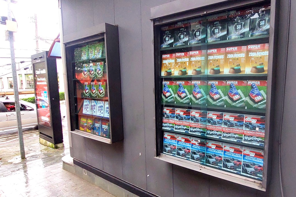 Duas vitrines, de uma banca de jornal, de vidro exibem revistas. A vitrine maior à direita mostra três edições da revista Super Interessante e um da Quatro Rodas. A banca menor à esquerda vê-se um Guia ENEM 2026 e Copas. O chão de cimento está molhado, e um carro prateado passa ao fundo.