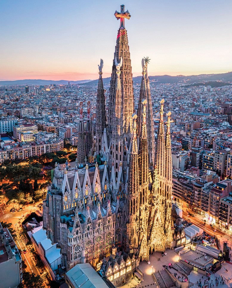 Vista aérea da Basílica da Sagrada Família em Barcelona, Espanha, ao entardecer. A arquitetura gótica da igreja é iluminada por luzes quentes, destacando suas torres pontiagudas e detalhes intrincados. Ao fundo, a cidade se estende com edifícios e ruas iluminadas, sob um céu em tons de azul e rosa.