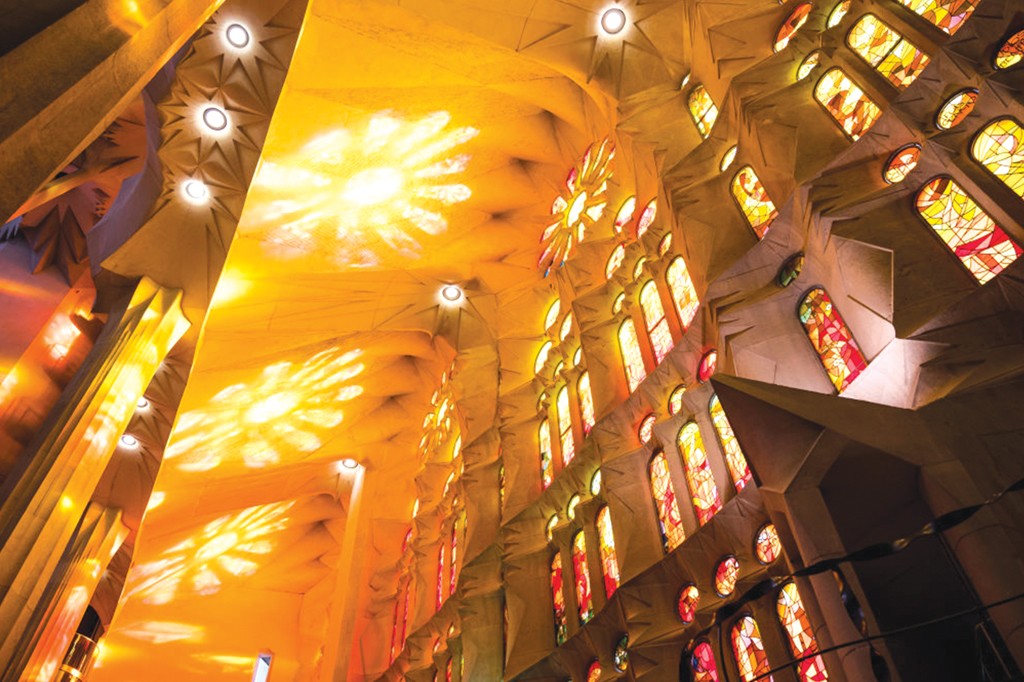 Interior de uma igreja com teto alto e abobadado, iluminado por luzes circulares e projeções de luz em formato de estrela. À direita, uma parede com múltiplos vitrais coloridos em tons de vermelho, laranja e amarelo, criando um ambiente quente e vibrante