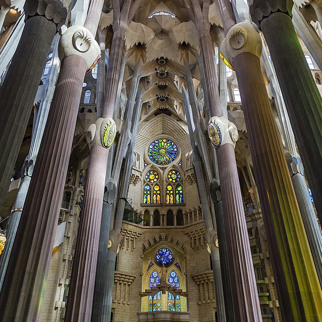 Interior da Basílica da Sagrada Família, com colunas altas e ramificadas que se assemelham a árvores, sustentando um teto abobadado. Há vitrais coloridos em formato circular e retangular, iluminando o ambiente com tons vibrantes de azul, verde, amarelo e vermelho. A arquitetura gótica modernista é grandiosa e detalhada