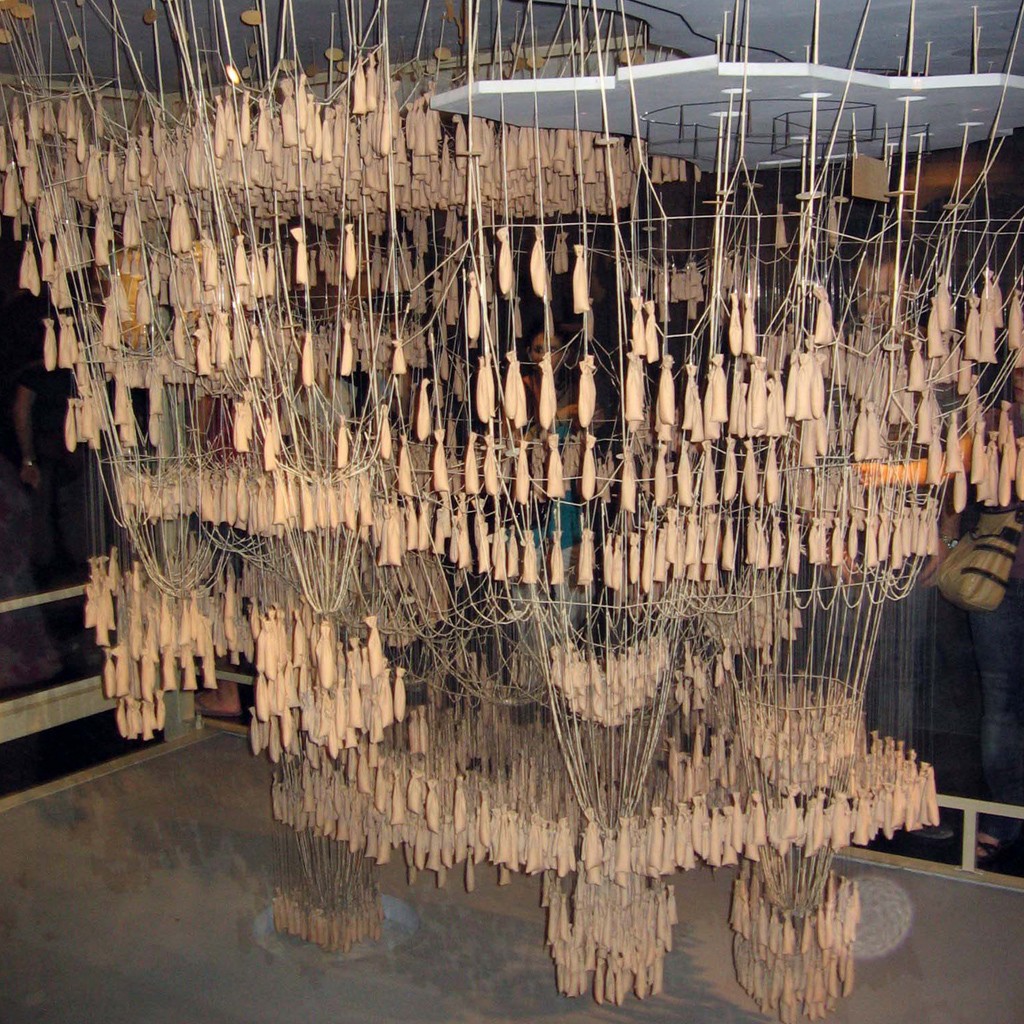 Instalação artística com centenas de pequenos sacos de tecido bege pendurados por fios finos, formando uma estrutura densa e orgânica que se estende do teto ao chão, com pessoas observando ao fundo