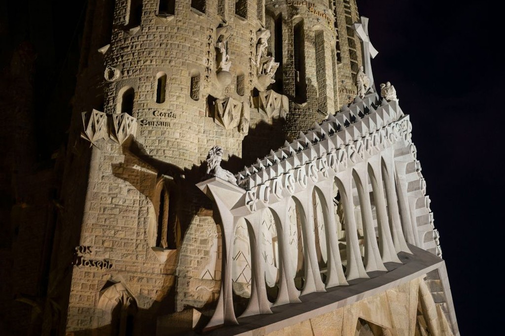 Fachada da Sagrada Família à noite, com iluminação destacando a torre de pedra e uma estrutura branca com arcos e pináculos triangulares. Esculturas e inscrições latinas como Corda Surcum e Sus Joseph são visíveis, contra um céu escuro
