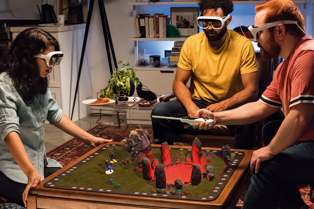 Três pessoas, duas sentadas e uma ajoelhada, usam óculos de realidade aumentada para jogar um jogo de tabuleiro com figuras holográficas de monstros e guerreiros. O homem ruivo à direita segura um controle branco, enquanto a mulher à esquerda e o homem de camiseta amarela observam o tabuleiro com atenção. O cenário é uma sala de estar com estantes e plantas.