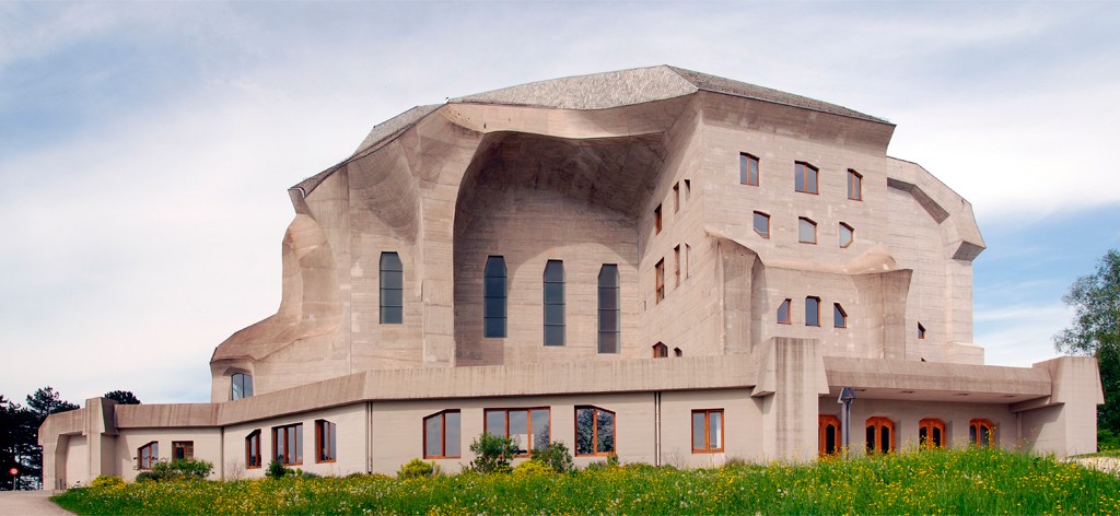 Edifício de concreto com arquitetura brutalista, formas orgânicas e telhado irregular, janelas retangulares e arqueadas, cercado por gramado verde com flores amarelas sob céu azul claro