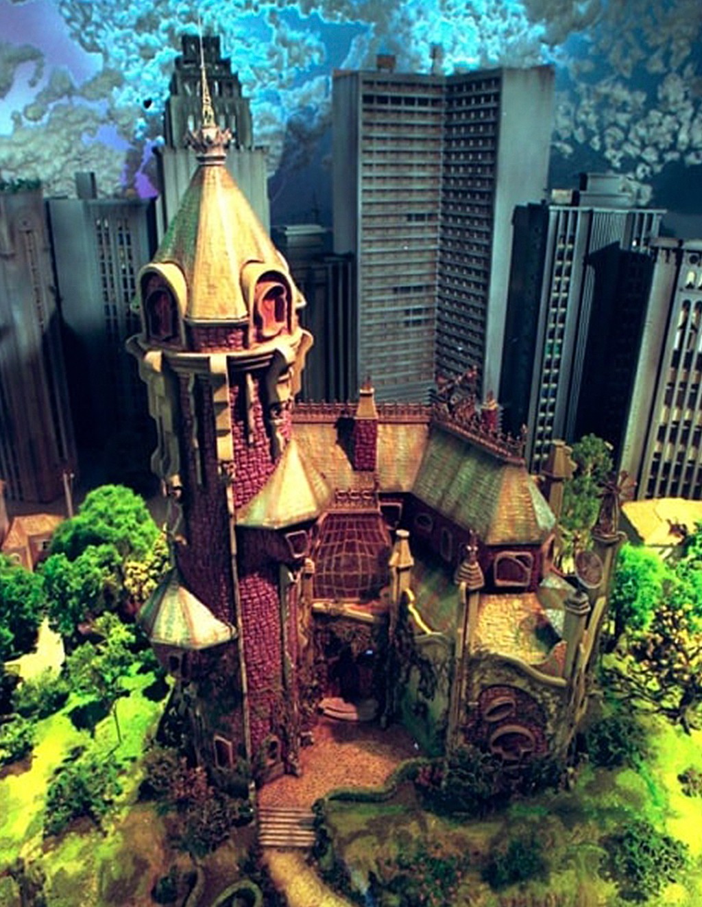 Maquete de um castelo gótico com telhados verdes e paredes avermelhadas, cercado por árvores e arbustos verdes, em contraste com arranha-céus cinzentos ao fundo sob um céu azul nublado