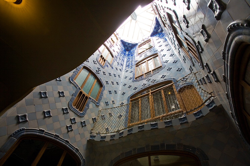 Vista de baixo para cima do pátio interno da Casa Batlló, com paredes curvas revestidas de azulejos azuis e brancos, janelas de madeira de formatos orgânicos e um telhado de vidro que ilumina o ambiente