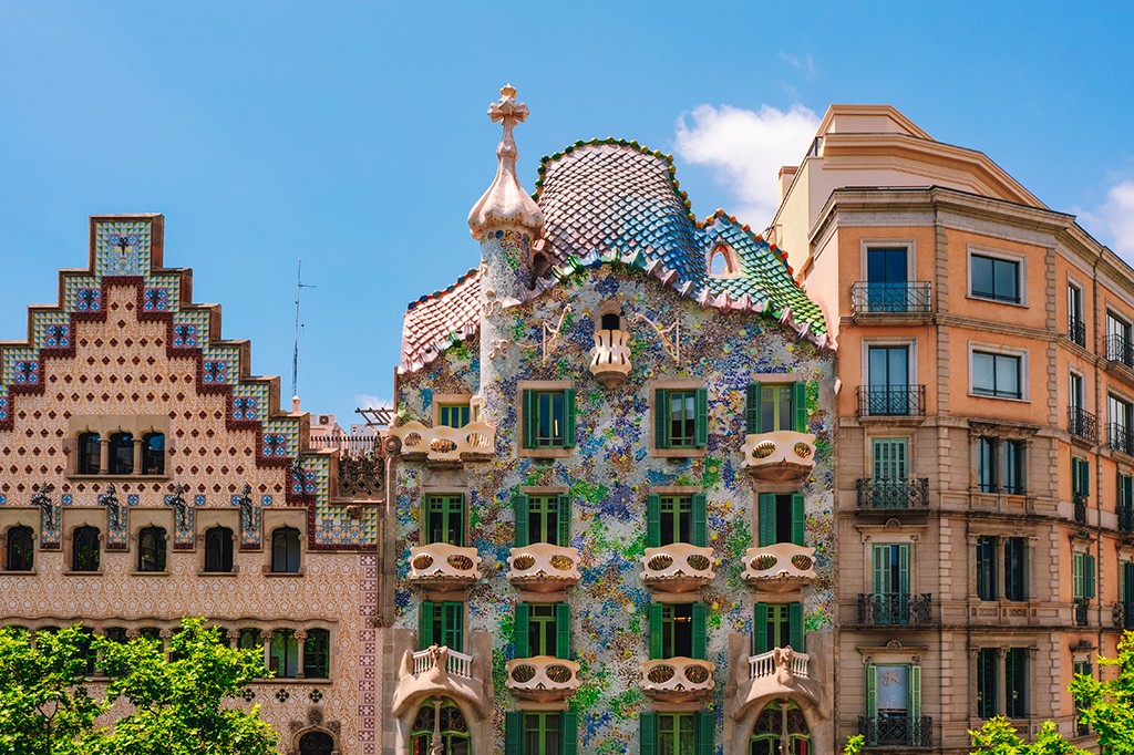 Três edifícios de arquitetura distinta sob céu azul. À esquerda, um prédio com fachada em tons de bege e marrom, com telhado em degraus e detalhes em mosaico azul. No centro, a Casa Batlló de Gaudí, com fachada colorida em mosaicos, telhado ondulado e chaminés decoradas. À direita, um edifício clássico de cor pêssego com sacadas de ferro forjado