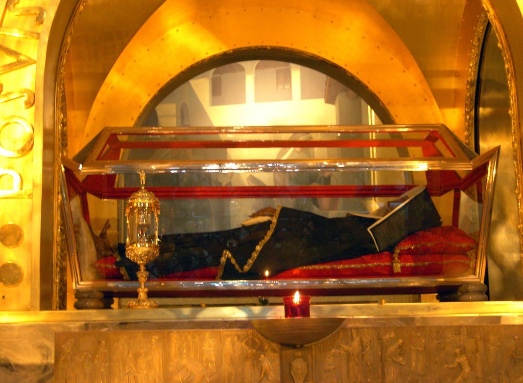 Corpo de Santa Rita de Cássia em um caixão de vidro e metal dourado, vestindo hábito escuro sobre um tecido vermelho, com uma vela acesa e um relicário dourado ao lado, em um ambiente iluminado por luz quente