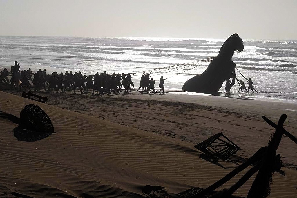 Silhuetas de dezenas de pessoas puxando cordas conectadas a uma grande estrutura escura em forma de cavalo, parcialmente submersa na água do mar, em uma praia arenosa com ondas ao fundo e céu claro