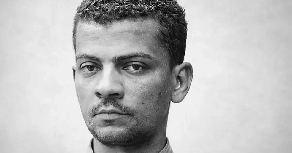 Homem de pele escura com cabelo crespo curto, barba rala e bigode, olhando diretamente para frente com expressão séria, em preto e branco