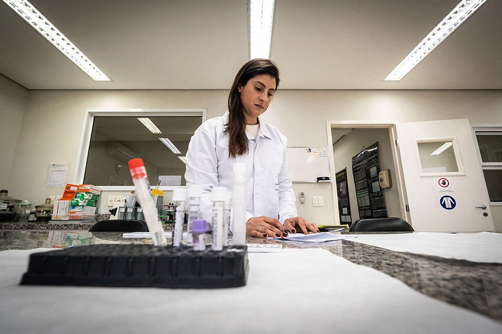 Mulher de jaleco branco, cabelos castanhos soltos, olhando para documentos sobre uma bancada de laboratório. Frascos e tubos de ensaio estão em primeiro plano.
