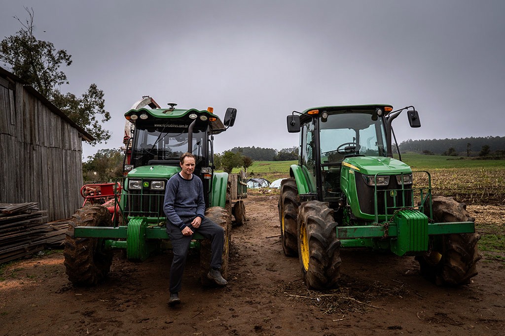 Homem de suéter azul e calça escura sentado no pneu de um trator verde, com outro trator similar ao lado, em um campo de terra batida sob céu nublado