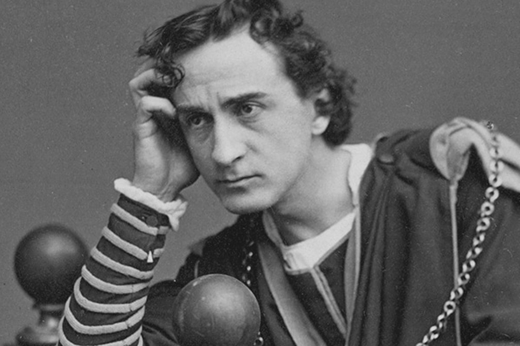 Homem com cabelo encaracolado, vestindo túnica escura e camisa listrada, apoia a cabeça na mão direita, olhando pensativo para a frente. Duas esferas escuras e uma corrente metálica aparecem no primeiro plano.