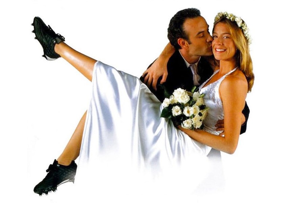 Um homem de terno preto beija a bochecha de uma mulher sorridente, vestida de noiva com um buquê de rosas brancas e uma coroa de flores. Ela está sendo carregada, com uma perna estendida para cima, calçando uma chuteira preta de futebol