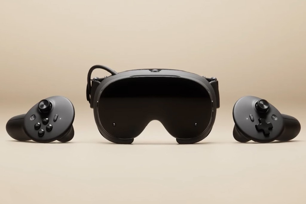 Um óculos de realidade virtual preto com lentes escuras e dois controles pretos, um de cada lado, sobre um fundo bege.